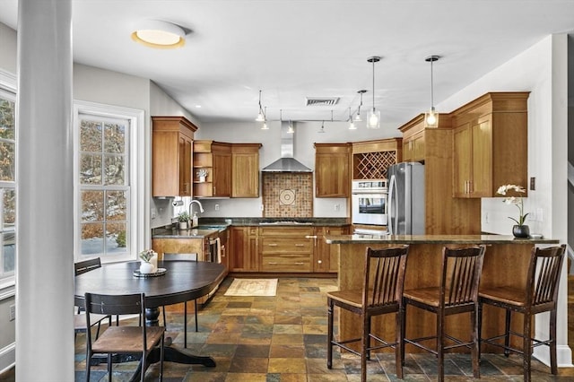 kitchen with pendant lighting, sink, appliances with stainless steel finishes, tasteful backsplash, and wall chimney exhaust hood