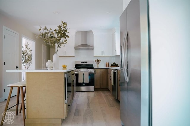 kitchen with appliances with stainless steel finishes, wall chimney range hood, light wood-style flooring, a kitchen bar, and tasteful backsplash