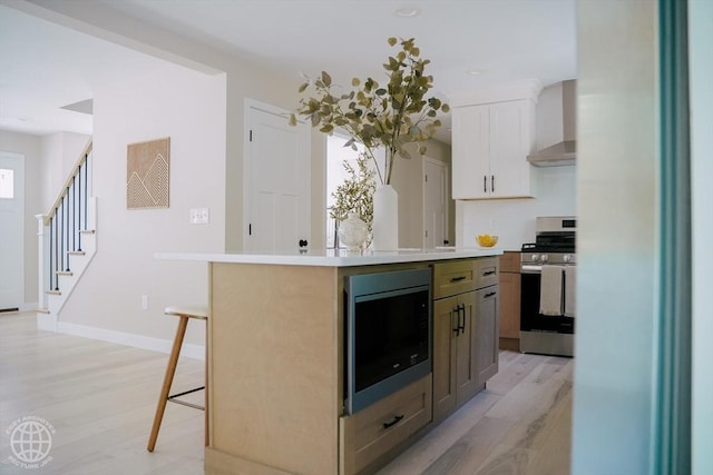 kitchen featuring stainless steel range with gas cooktop, built in microwave, wall chimney exhaust hood, light wood-style flooring, and a breakfast bar