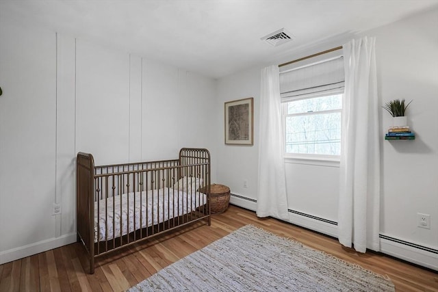 bedroom with a nursery area, baseboard heating, wood finished floors, and visible vents