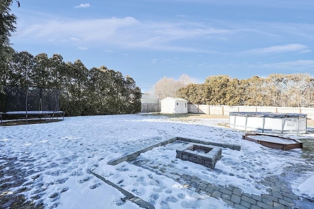 yard layered in snow featuring an outdoor fire pit, a fenced backyard, a fenced in pool, a storage unit, and a trampoline