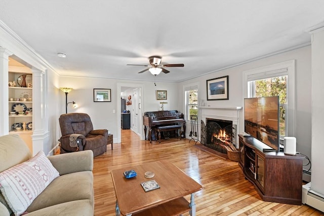 living area featuring a ceiling fan, a baseboard radiator, a warm lit fireplace, ornamental molding, and ornate columns