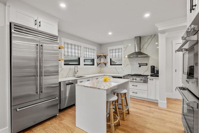 kitchen with light wood finished floors, stainless steel appliances, white cabinetry, wall chimney exhaust hood, and a sink