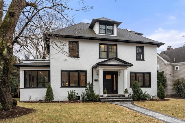 traditional style home featuring a shingled roof, a front lawn, a balcony, and stucco siding