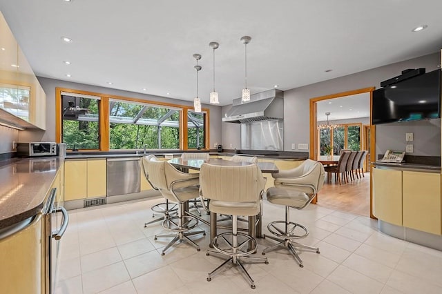 kitchen with hanging light fixtures, stainless steel dishwasher, light tile patterned floors, and wall chimney exhaust hood