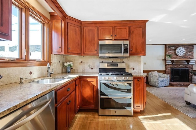 kitchen featuring stainless steel appliances, light wood-style flooring, a brick fireplace, a sink, and light stone countertops