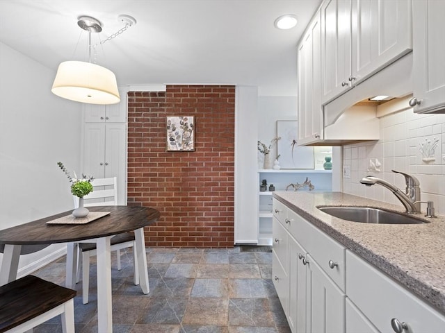 kitchen with brick wall, pendant lighting, white cabinets, light stone counters, and sink