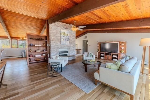 living area with ceiling fan, vaulted ceiling with beams, a stone fireplace, wood finished floors, and a baseboard radiator
