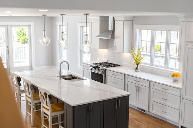 kitchen featuring an island with sink, wall chimney range hood, white cabinetry, a sink, and stainless steel range