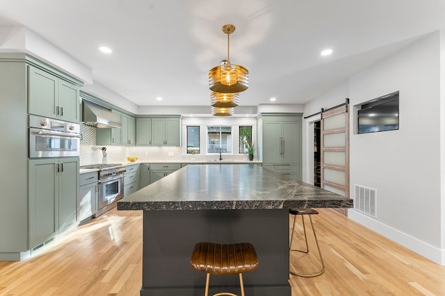 kitchen featuring wall chimney exhaust hood, stainless steel appliances, a breakfast bar area, green cabinets, and a barn door