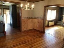 unfurnished dining area featuring a notable chandelier, wood walls, wainscoting, radiator heating unit, and dark wood finished floors