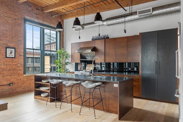 kitchen featuring light hardwood / wood-style flooring, brick wall, an island with sink, a towering ceiling, and beamed ceiling