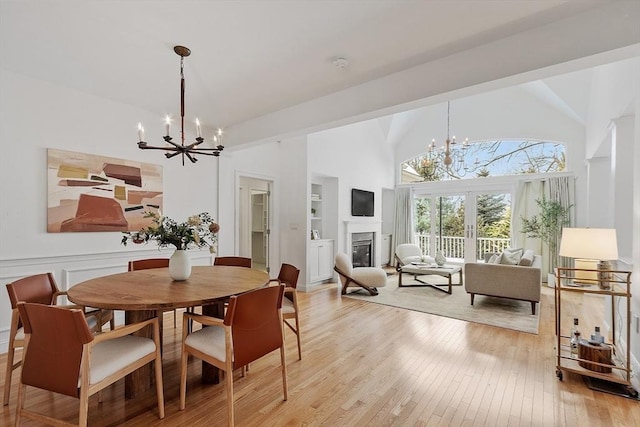 dining room with built in shelves, light wood finished floors, an inviting chandelier, a glass covered fireplace, and high vaulted ceiling