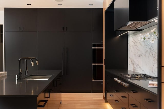 kitchen featuring sink, wall chimney range hood, backsplash, stainless steel gas stovetop, and light wood-type flooring