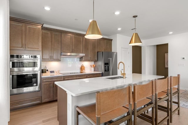 kitchen with under cabinet range hood, a sink, light wood-style floors, decorative backsplash, and appliances with stainless steel finishes