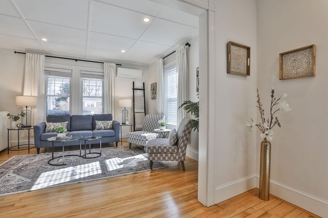 living room with a paneled ceiling, hardwood / wood-style floors, baseboards, and an AC wall unit