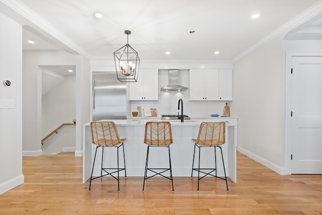 kitchen with wall chimney range hood, stainless steel built in refrigerator, light wood-type flooring, white cabinets, and ornamental molding