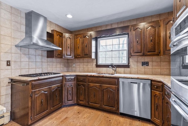 kitchen with a sink, stainless steel appliances, wall chimney exhaust hood, light countertops, and light wood-style floors