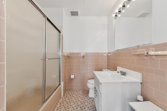 bathroom featuring tile walls, toilet, vanity, shower / bath combination with glass door, and a wainscoted wall