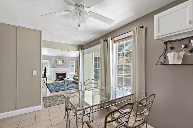 dining space with baseboards, a ceiling fan, a warm lit fireplace, and light tile patterned flooring