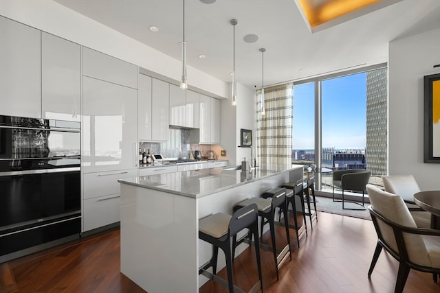 kitchen featuring white cabinets, a center island with sink, decorative backsplash, multiple ovens, and dark hardwood / wood-style flooring