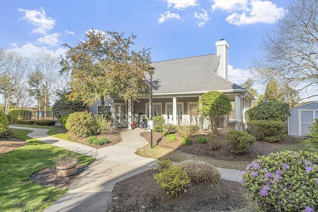 view of front of house featuring a shingled roof, an outbuilding, covered porch, a chimney, and a storage unit