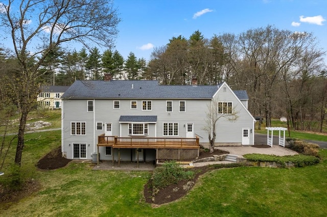 rear view of house featuring a shingled roof, a deck, a chimney, a lawn, and a patio