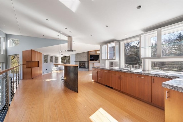 kitchen with a large fireplace, open floor plan, wall chimney range hood, light wood-type flooring, and brown cabinets