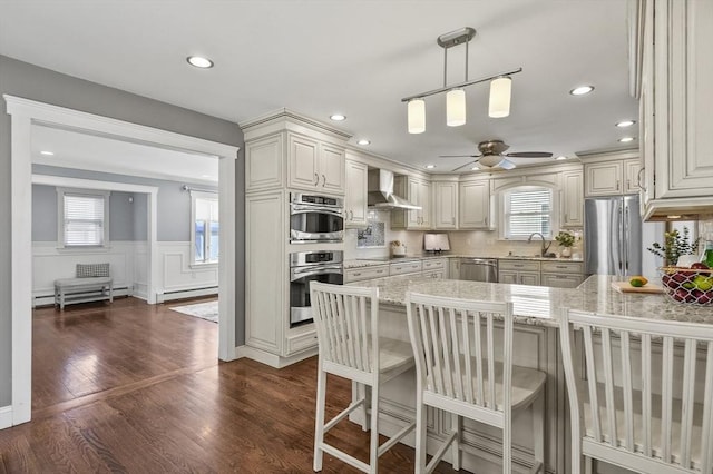 kitchen with wall chimney exhaust hood, hanging light fixtures, appliances with stainless steel finishes, ceiling fan, and sink