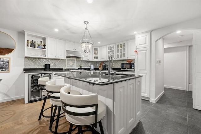 kitchen featuring beverage cooler, dark countertops, stainless steel microwave, under cabinet range hood, and a kitchen island with sink
