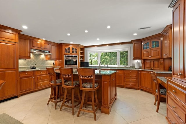 100 Parmenter Rd, Framingham, MA 01701 - kitchen featuring stainless steel oven, under cabinet range hood, backsplash, brown cabinetry, and glass insert cabinets