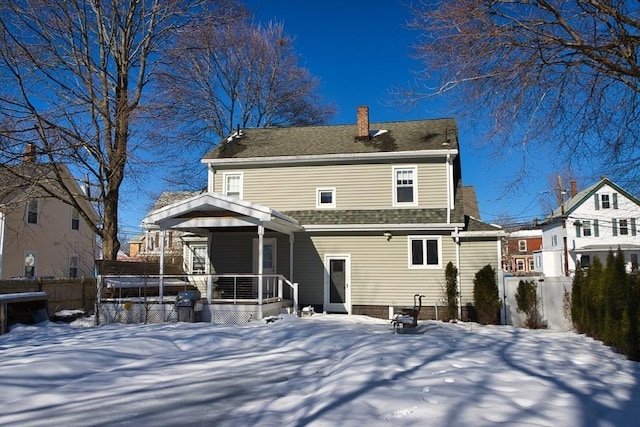 snow covered house featuring a shingled roof, a chimney, fence, and a porch