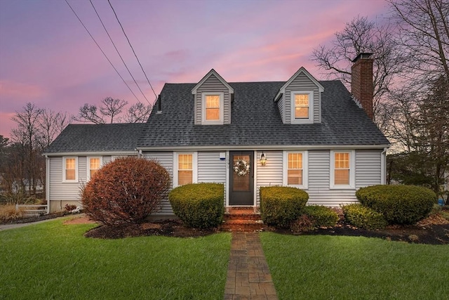 cape cod house with a chimney, a yard, and roof with shingles