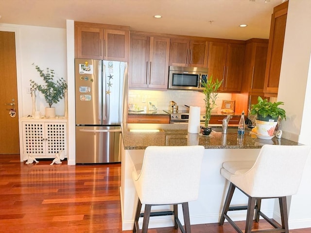 kitchen featuring stainless steel appliances, brown cabinets, a breakfast bar area, decorative backsplash, and dark stone counters