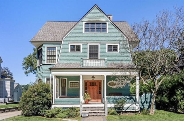 shingle-style home with a porch, a chimney, a balcony, and roof with shingles