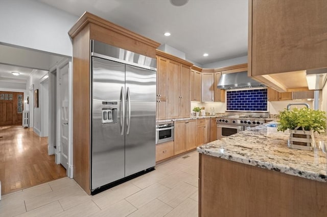 kitchen with light stone counters, light brown cabinets, appliances with stainless steel finishes, and wall chimney exhaust hood