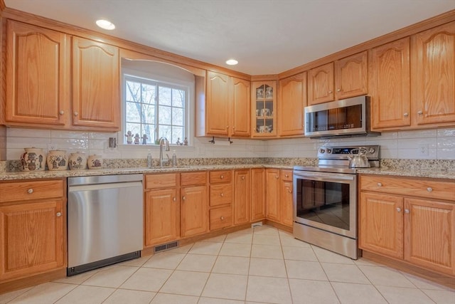 kitchen featuring stainless steel appliances, backsplash, a sink, light stone countertops, and glass insert cabinets