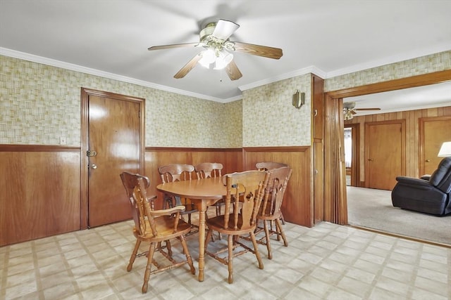 dining room with light flooring, a ceiling fan, wainscoting, wallpapered walls, and wood walls