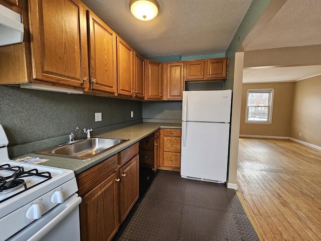 kitchen with brown cabinetry, a sink, a textured ceiling, white appliances, and baseboards