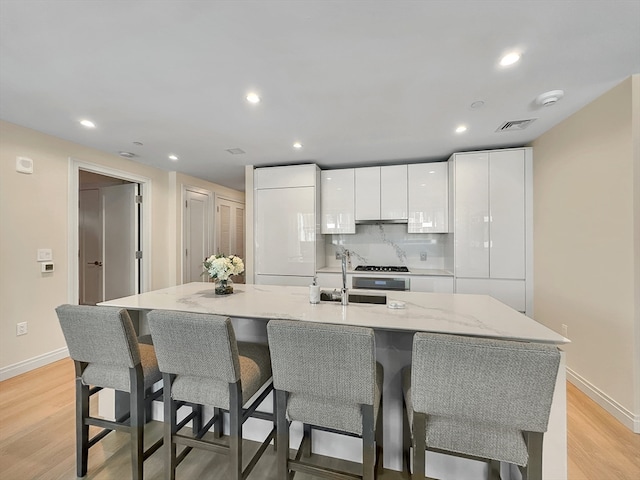 kitchen with white cabinetry, decorative backsplash, light wood-type flooring, light stone countertops, and a center island with sink