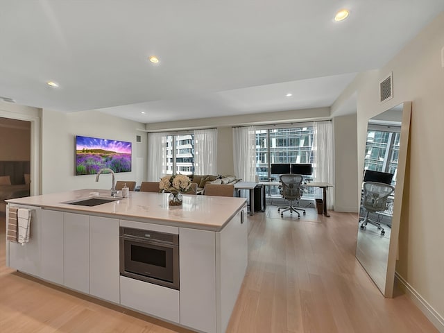 kitchen with stainless steel oven, white cabinets, an island with sink, light wood-type flooring, and sink