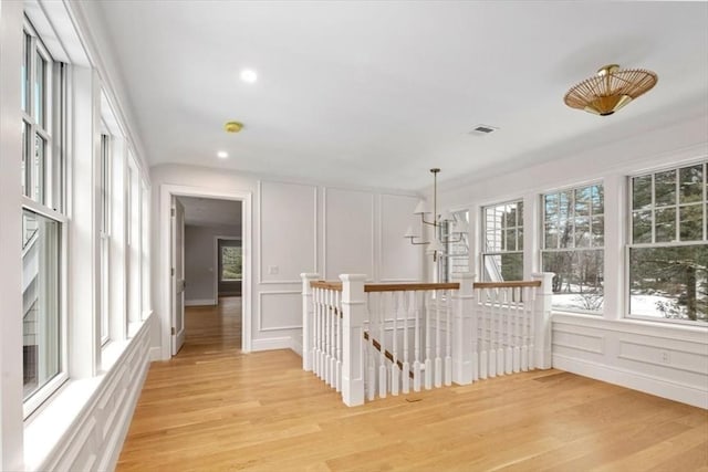 corridor featuring light wood-style flooring, visible vents, a decorative wall, and an upstairs landing