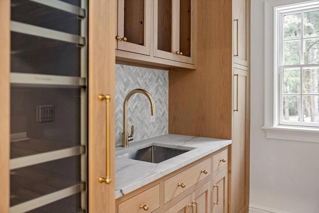 kitchen featuring decorative backsplash, light brown cabinetry, glass insert cabinets, a sink, and light stone countertops