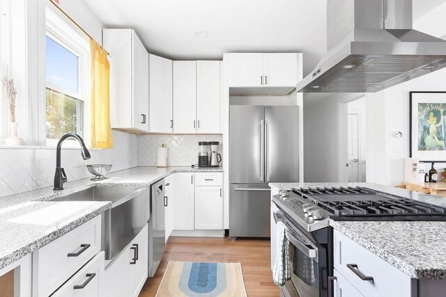 kitchen featuring wall chimney range hood, stainless steel appliances, a sink, light stone countertops, and light wood-style flooring