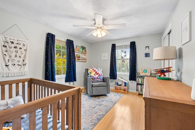 bedroom with multiple windows, light wood-style floors, a nursery area, and a ceiling fan