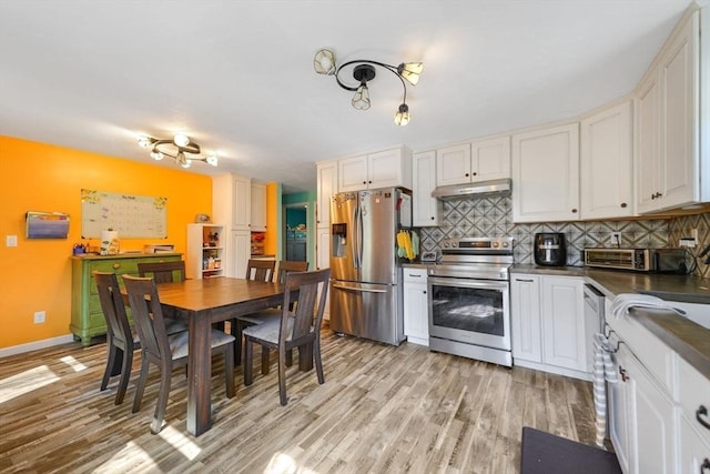 kitchen featuring dark countertops, light wood-style floors, under cabinet range hood, appliances with stainless steel finishes, and backsplash