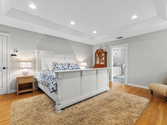 bedroom with a tray ceiling, light wood-style flooring, baseboards, recessed lighting, and ensuite bath