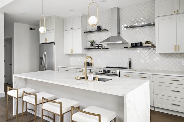 kitchen with an island with sink, appliances with stainless steel finishes, wall chimney range hood, and decorative light fixtures