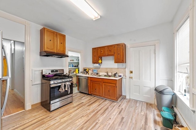 kitchen with light wood-style flooring, appliances with stainless steel finishes, brown cabinetry, a sink, and under cabinet range hood