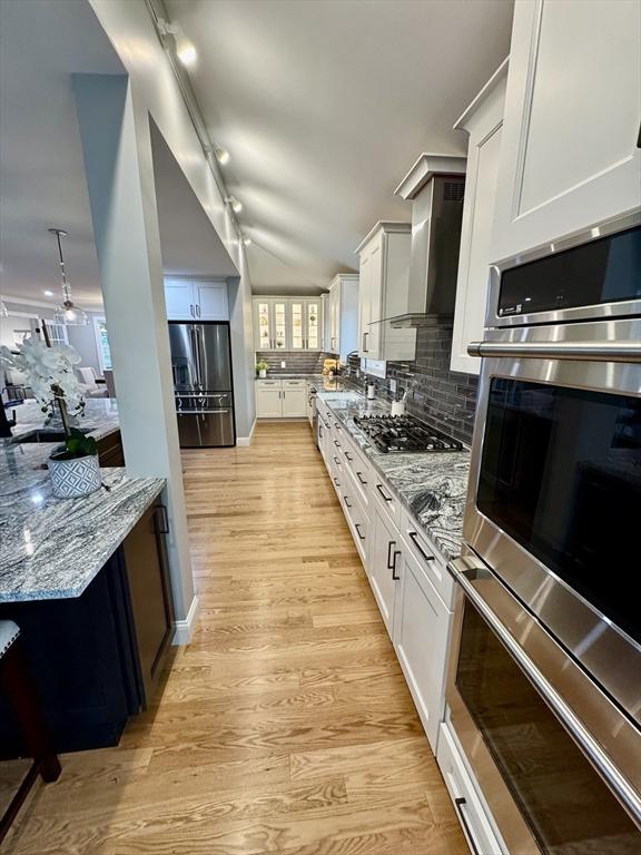kitchen with appliances with stainless steel finishes, wall chimney exhaust hood, light wood-style flooring, white cabinetry, and vaulted ceiling
